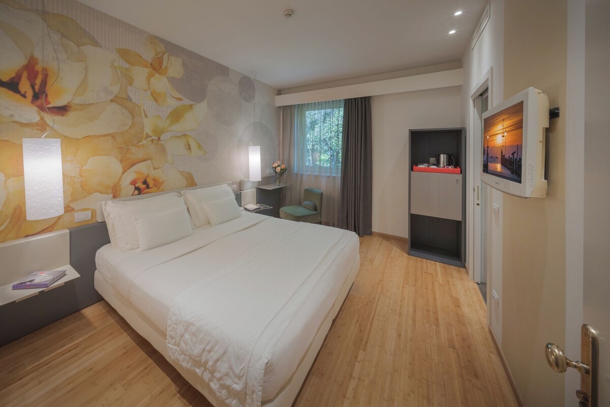 Habitación de hotel con una cama grande, un televisor montado en la pared, una mesita de noche con lámpara y una ventana con cortinas.