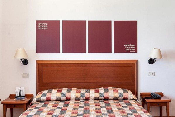 A hotel room with a wooden headboard, a quilted bedspread, two bedside tables, a wall-mounted lamp, and a framed artwork above the bed. - Il Brigantino (Porto Recanati)