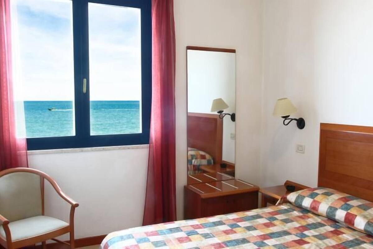 Een hotelkamer met een bed, een stoel, een nachtkastje, een spiegel en een raam met uitzicht op zee.