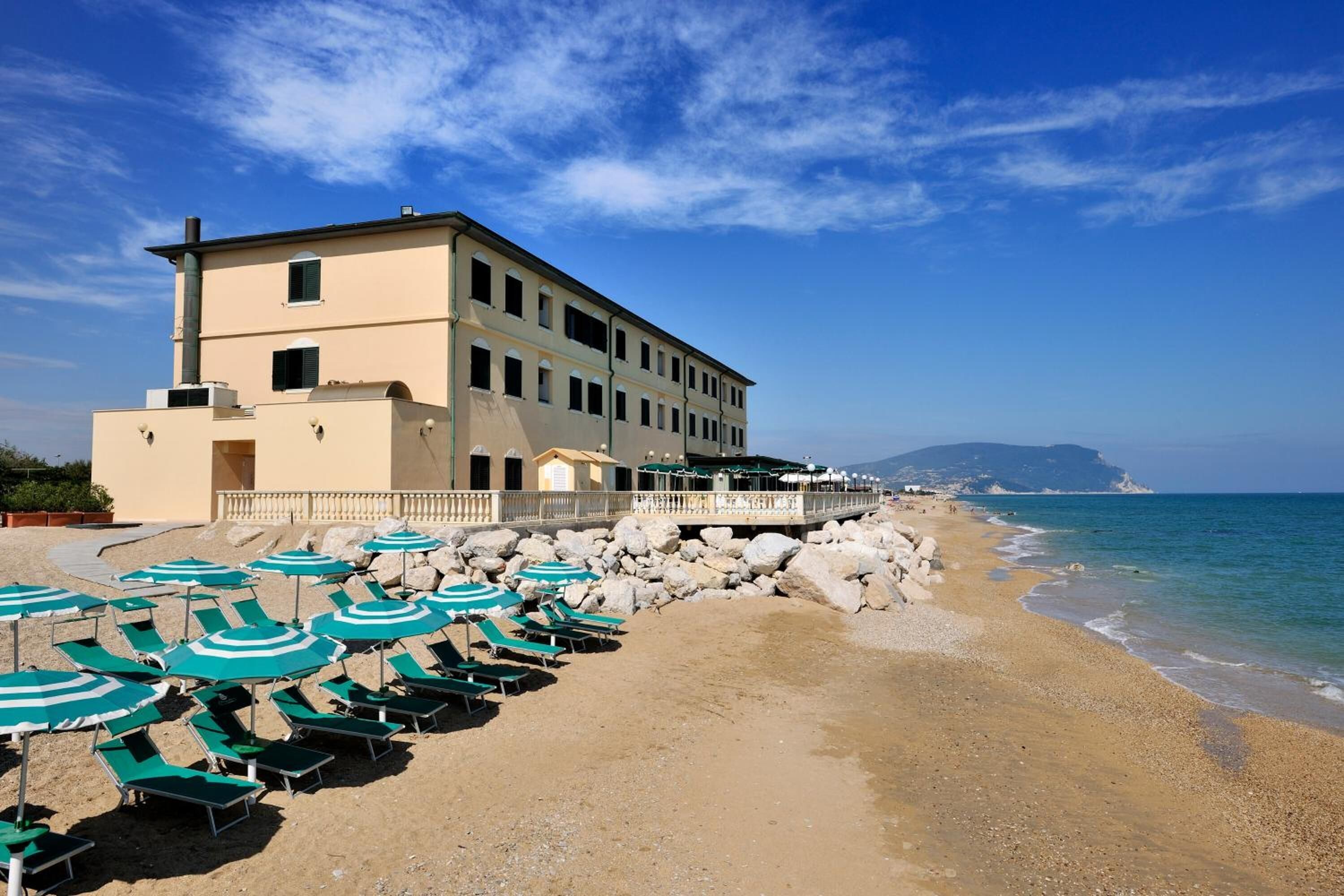 Un hotel frente a la playa con numerosas sillas de descanso y sombrillas, un edificio grande con balcón y vistas al mar y colinas distantes.