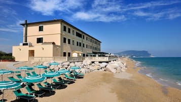 Un hotel frente a la playa con numerosas sillas de descanso y sombrillas, un edificio grande con balcón y vistas al mar y colinas distantes.