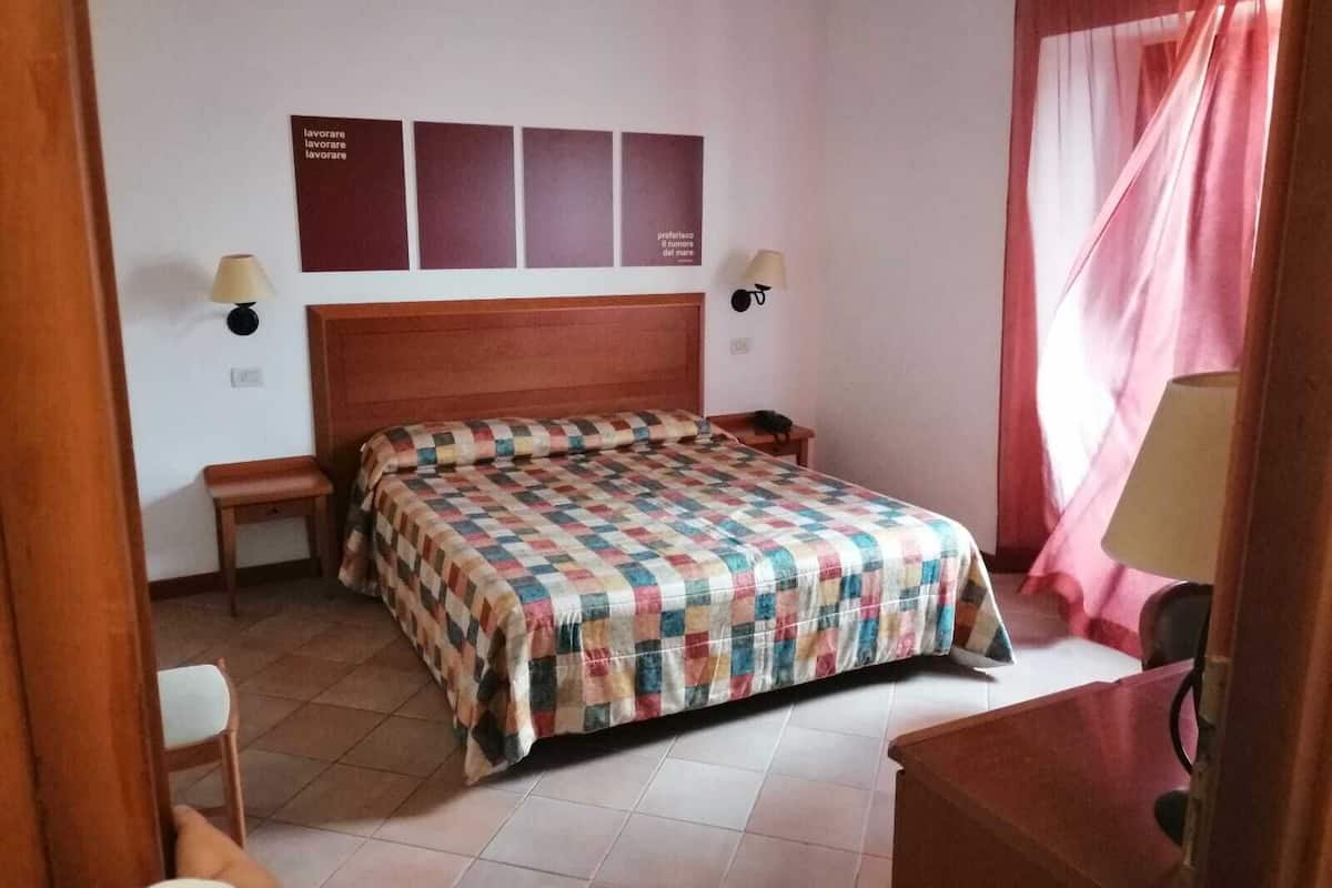 Een slaapkamer met een houten hoofdeinde, een geruit bedlaken, twee nachtkastjes met lampen, een klein houten bankje en een bureau met stoel.