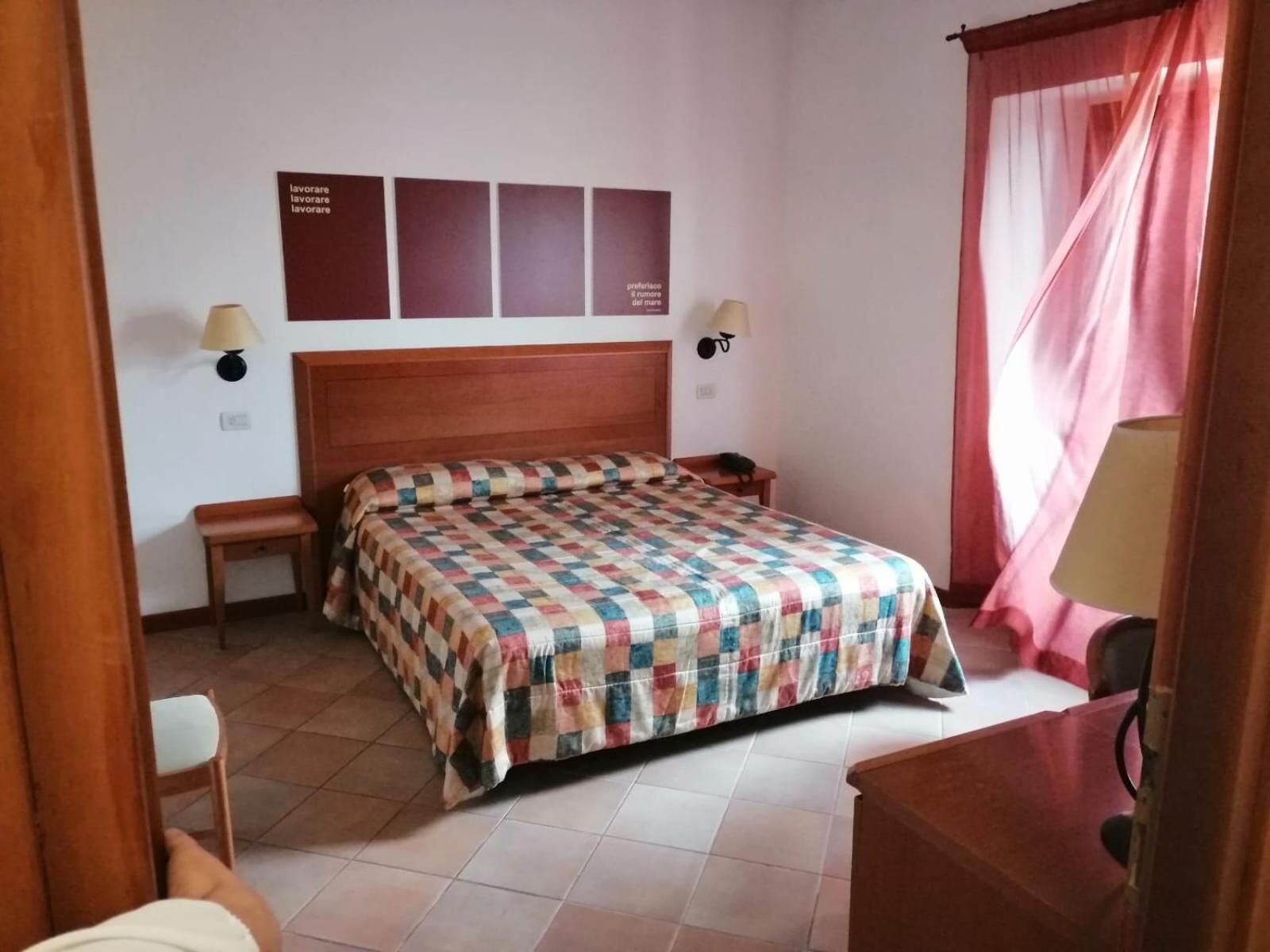 Habitación de hotel con cama de madera, un banco, un escritorio y una silla. Hay dos lámparas de pared y un reloj de pared.
