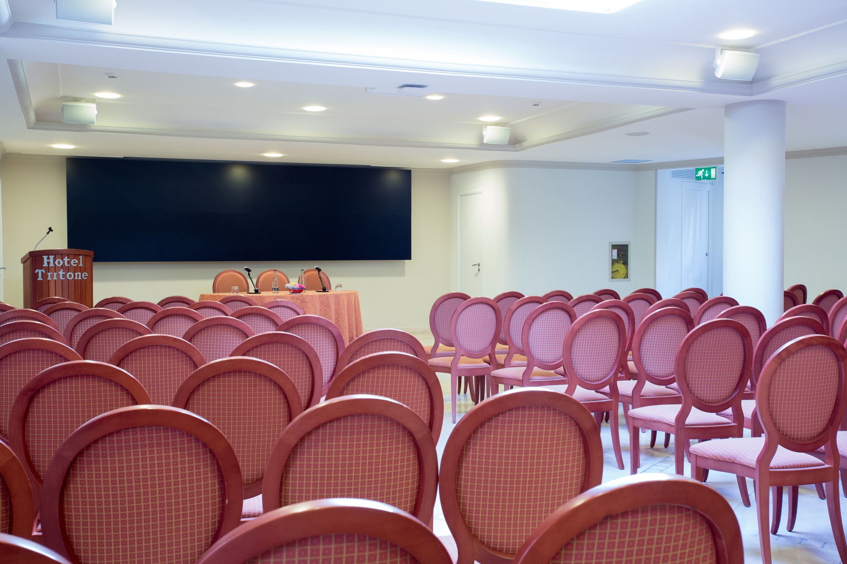 Uma sala de conferências com fileiras de cadeiras, um púlpito com a inscrição 'Hotel Tritone' e uma tela grande.