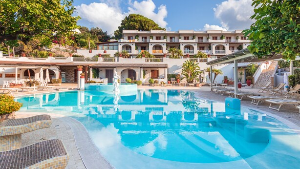 A swimming pool with lounge chairs and a building with multiple balconies in the background. - Hotel Tritone (Lipari)