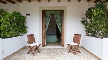 Uma varanda com duas cadeiras de madeira, piso de azulejos e uma porta com cortinas verdes.