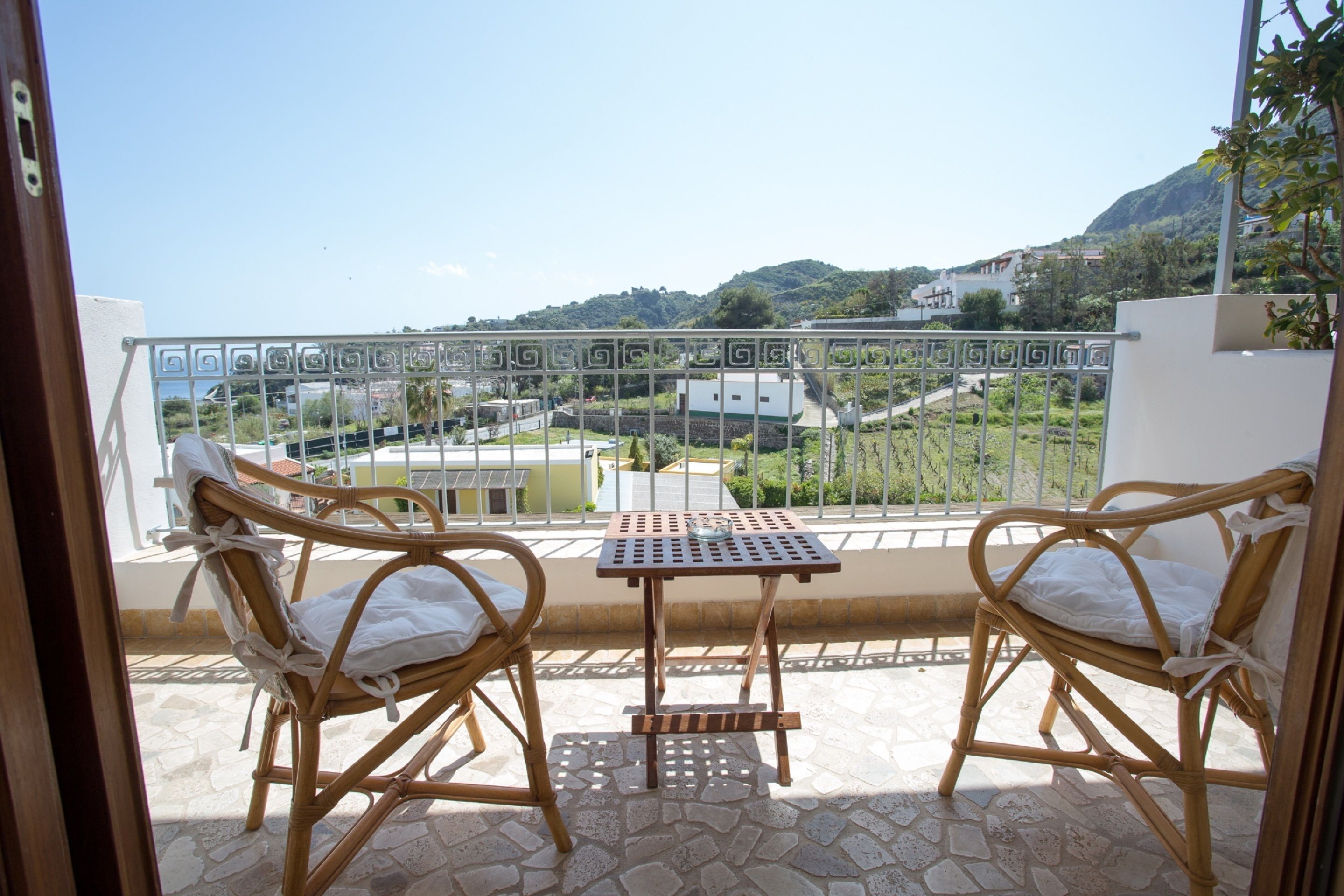 A balcony with wicker chairs and a small table, overlooking a residential area with houses and greenery.
