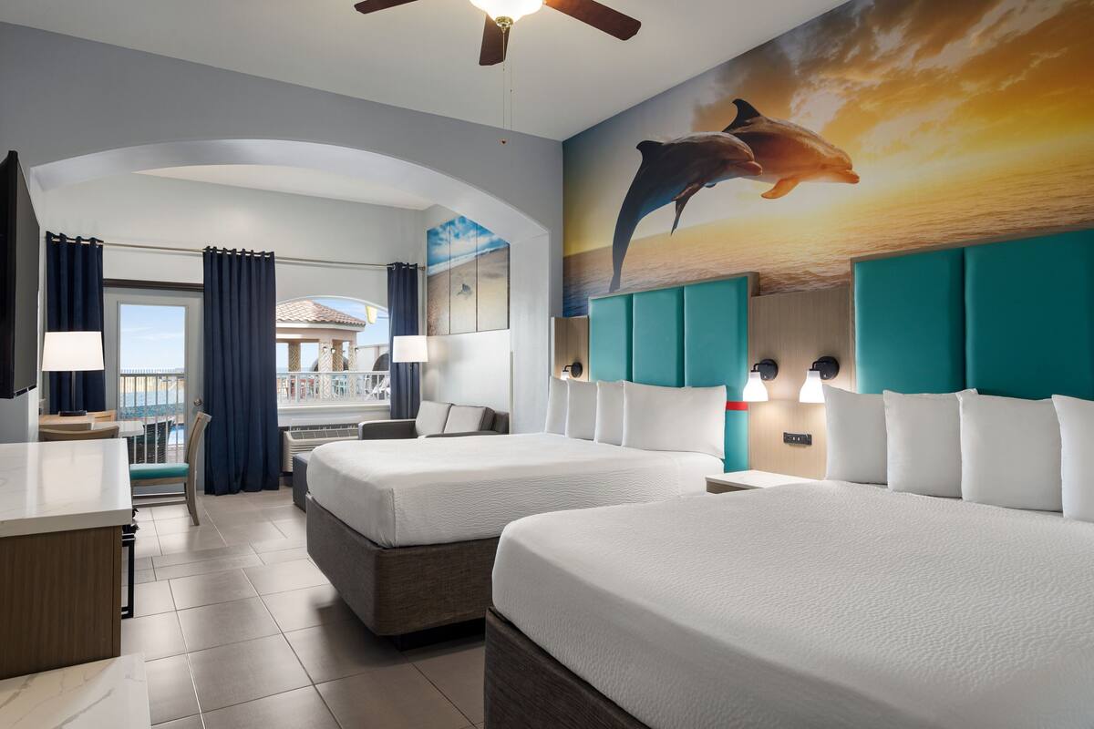 Habitación de hotel con dos camas, un escritorio y vistas al balcón.