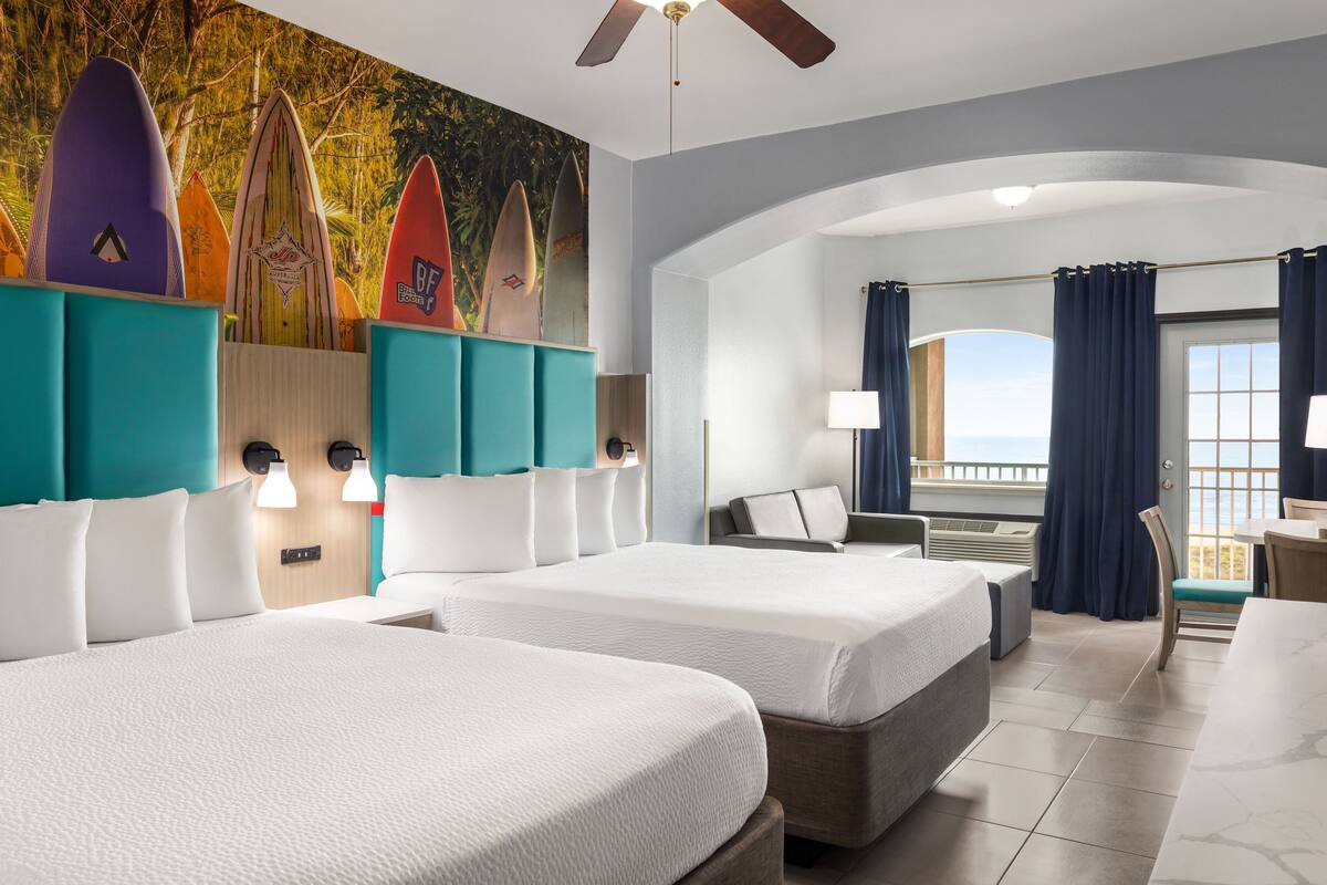 Habitación de hotel con dos camas, un ventilador de techo, un mural de tablas de surf y vistas al océano.