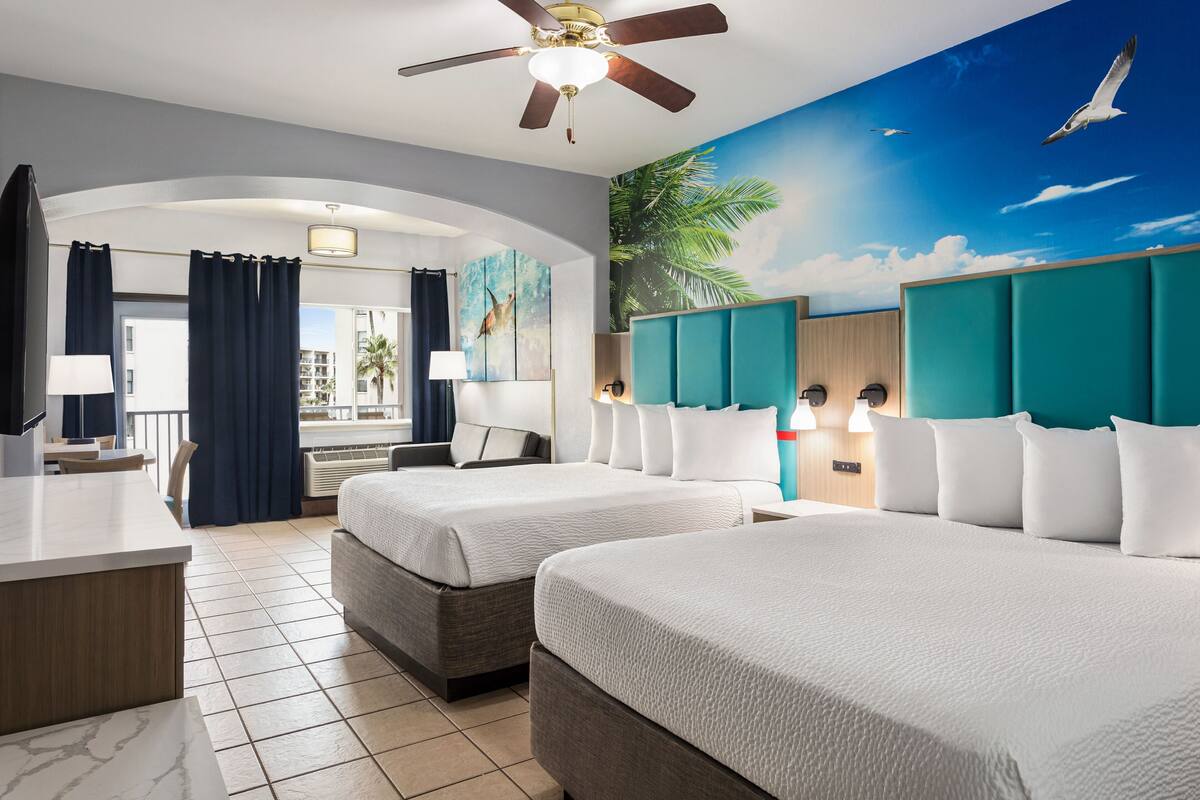 Habitación de hotel con dos camas, un escritorio, un ventilador de techo y un mural con una escena tropical.