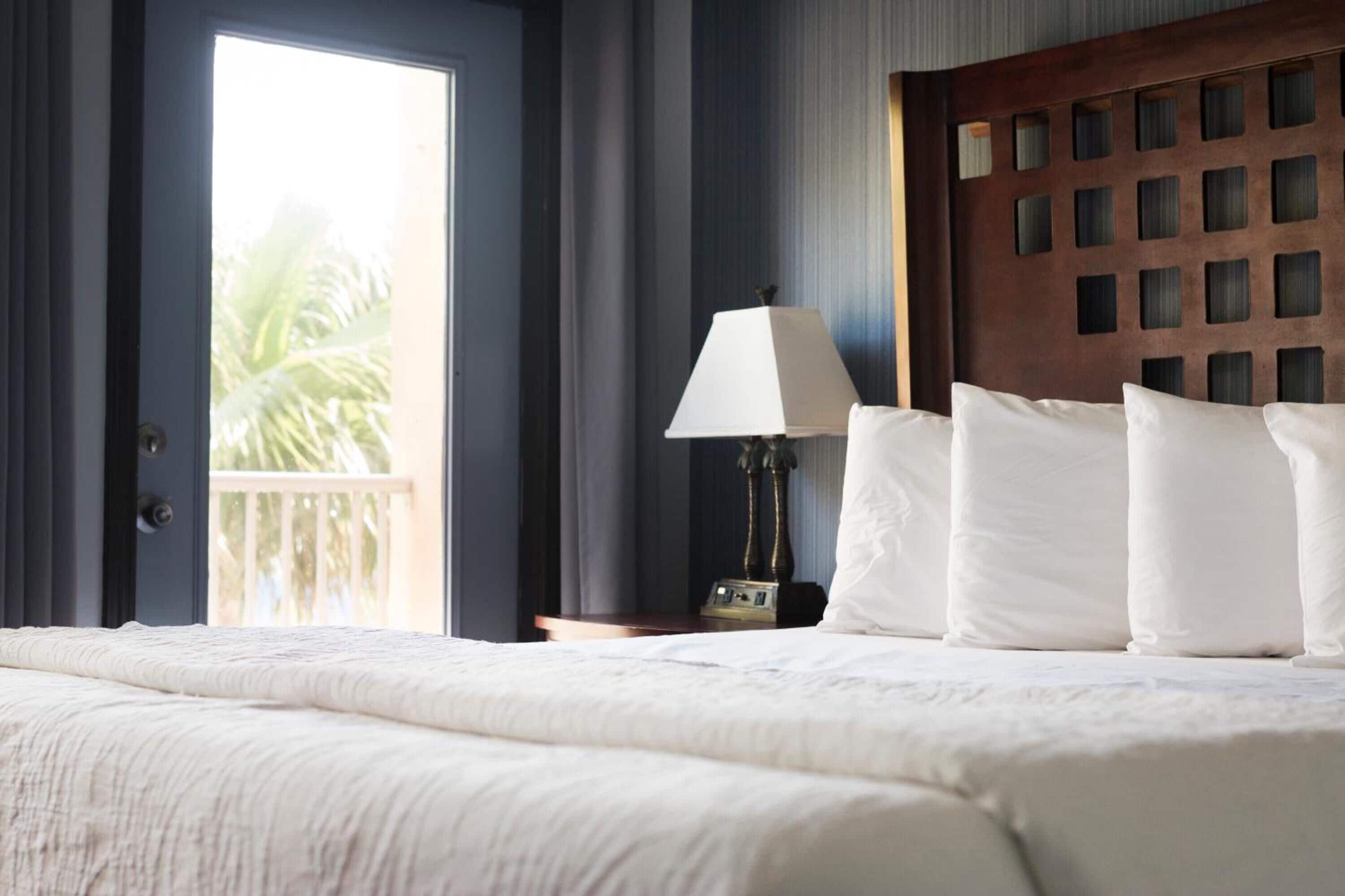 Habitación de hotel con una cama grande, una mesita de noche con lámpara y vistas a palmeras a través de una puerta corrediza.