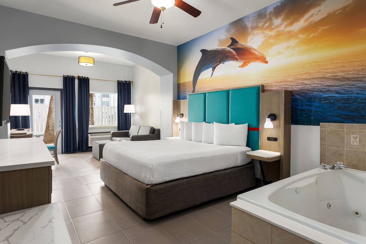 Una habitación de hotel moderna con una cama grande, zona de estar, escritorio, baño con bañera y un mural de delfines en la pared.