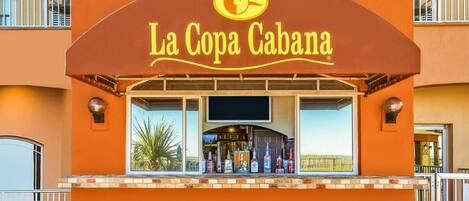 A bar named La Copa Cabana with a large sign, a window display of bottles, and a view of the landscape.