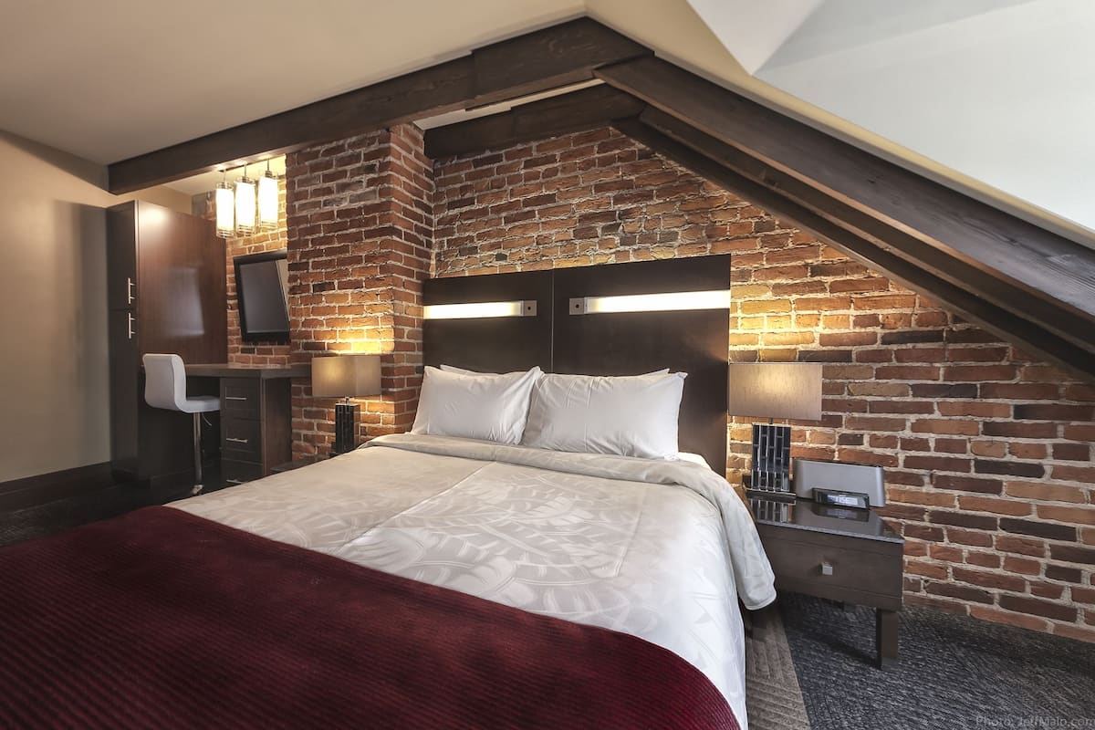 Une chambre avec un mur en briques, un lit recouvert de draps blancs, une table de chevet avec une lampe et un bureau avec une chaise.