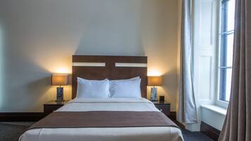 Una camera d'albergo con un letto grande, due comodini con lampade, una finestra con tende e una testiera con illuminazione.