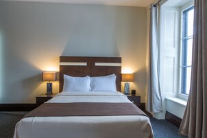 Una camera d'albergo con un letto grande, due comodini con lampade, una finestra con tende e una testiera con illuminazione.