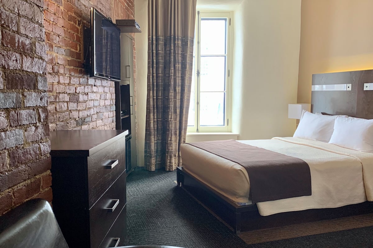 Une chambre d’hôtel avec un mur en briques, un lit recouvert de draps blancs, une commode en bois foncé et une fenêtre avec des rideaux.