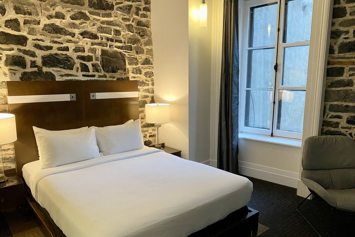 Un lit bien fait, avec du linge de lit blanc, dans une pièce dont un mur est en pierre et qui possède une fenêtre avec des rideaux.