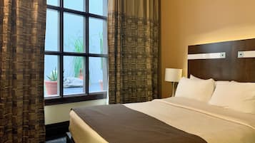 A hotel room with a large bed, a window with curtains, a nightstand with a lamp, and a view of the outside.
