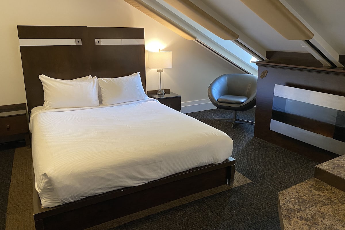 Un lit bien fait, avec du linge de lit blanc, dans une chambre d’hôtel dotée d’un plafond en pente et d’une chaise moderne.