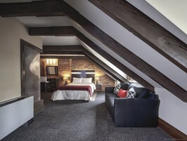 Un dormitorio con techo inclinado, pared de ladrillo, cama, mesita de noche, lámpara y sofá.
