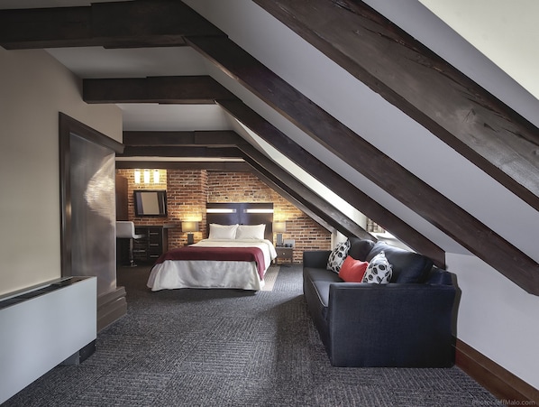 A bedroom with a sloped ceiling, a brick wall, a bed, a nightstand, a lamp, and a sofa.