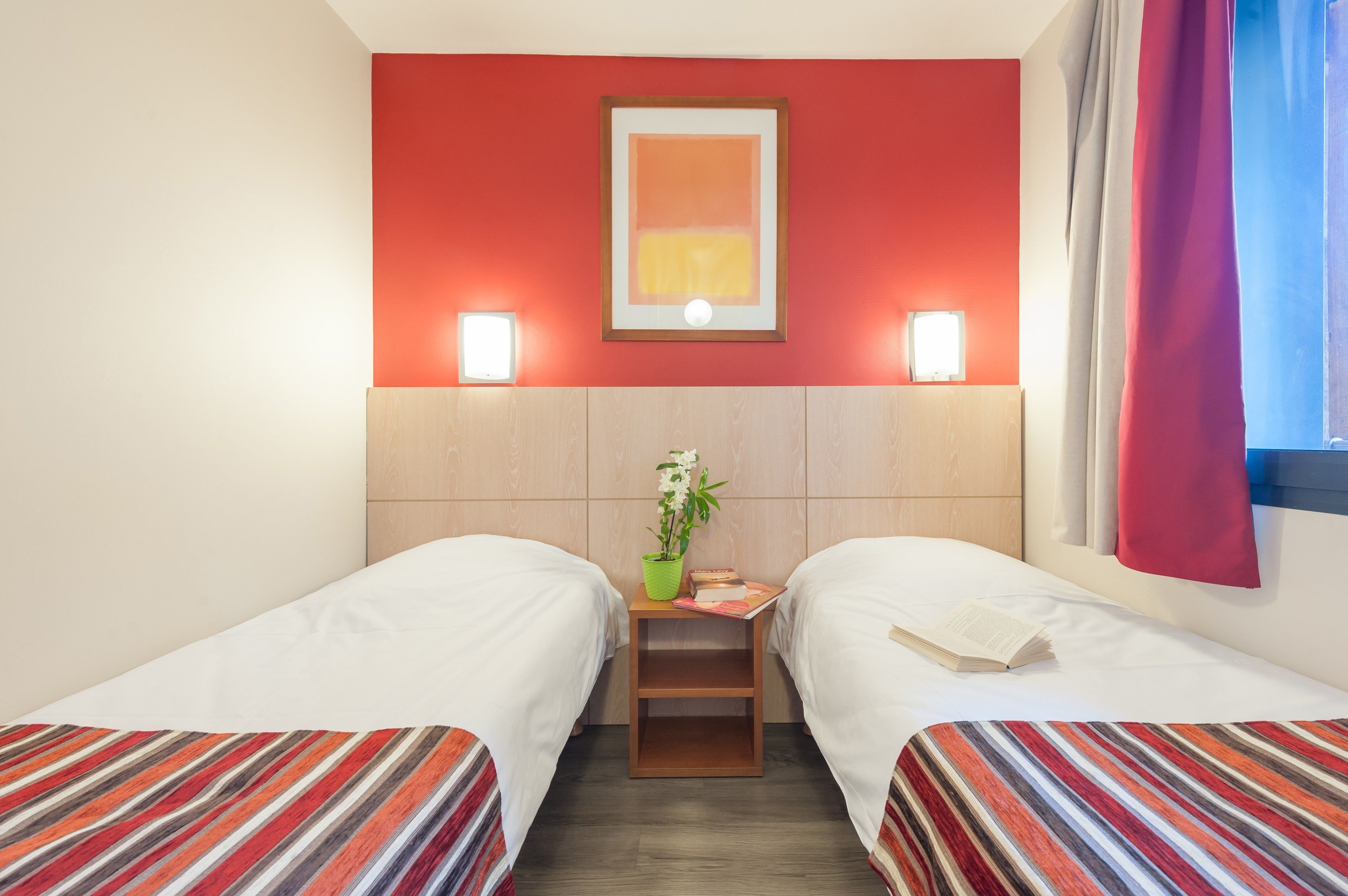 A hotel room with two single beds, a small table with a plant, a framed picture on the wall, and two wall-mounted lights.