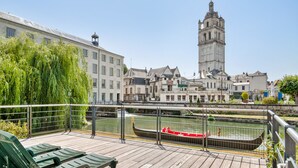 Una terraza de madera con sillas verdes, una góndola en un canal y edificios históricos al fondo.