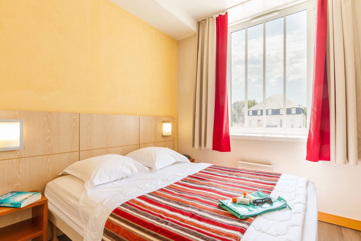 A hotel room with a bed, a nightstand, a window with curtains, and a view of buildings outside.