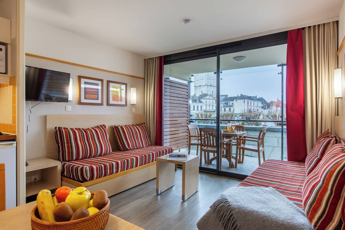 A modern hotel room with a sofa, a dining table, and a large window with a view of buildings.