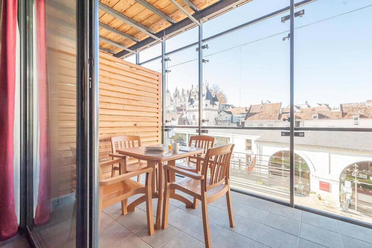 A balcony with a wooden table and chairs, large windows offering a view of a historic building.