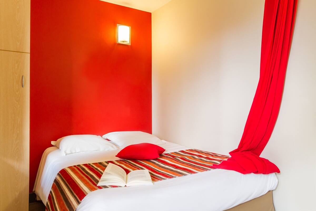 A single bed with a striped bedspread, white pillows, and a red pillow. There is a book on the bed.