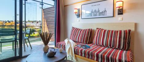 A hotel room with a large window, a sofa with striped cushions, a small table with a teapot and a plate of pastries, and a framed picture on the wall.