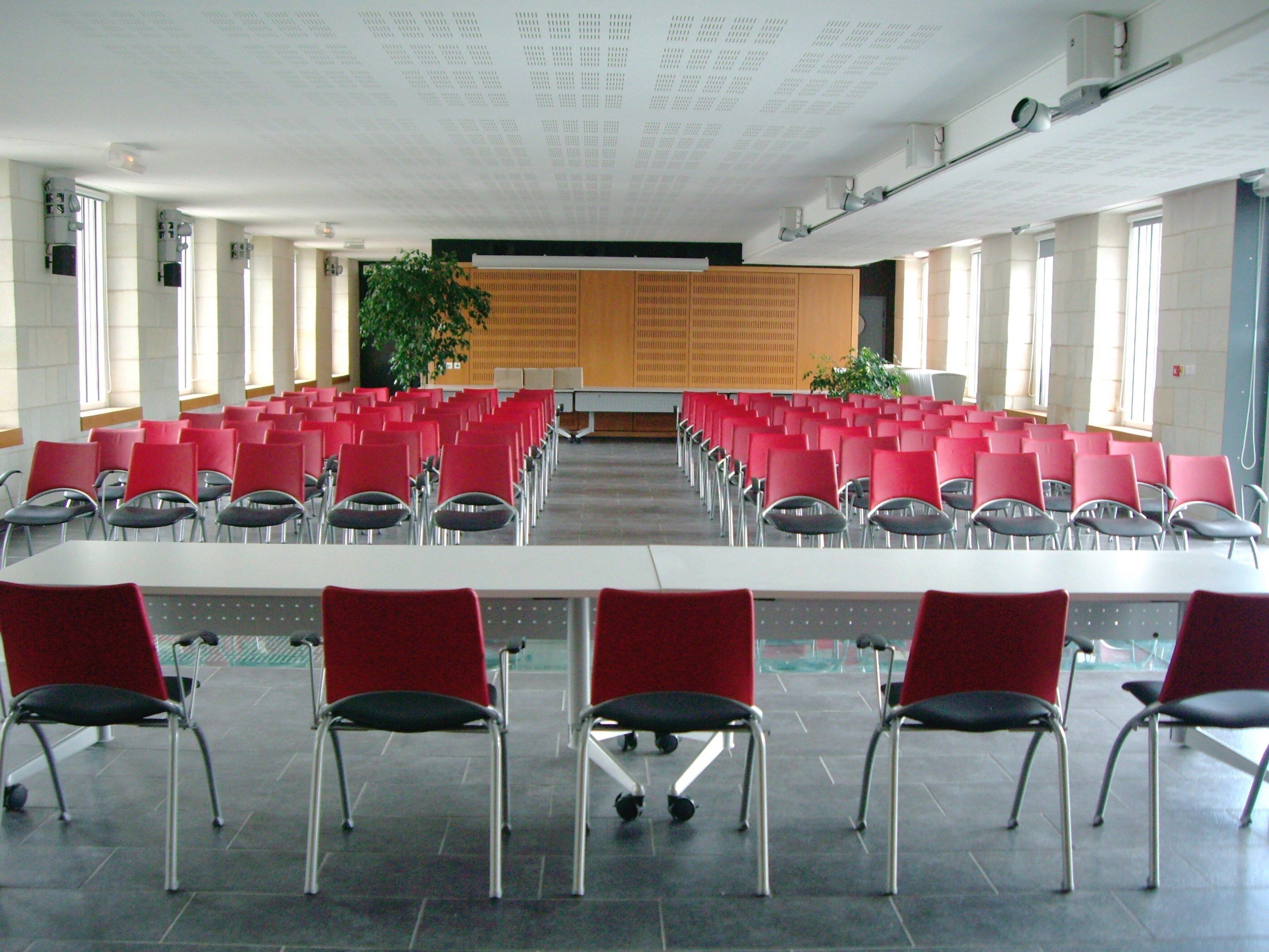 A conference room with rows of red chairs and a long table, large windows, and a ceiling with recessed lighting.
