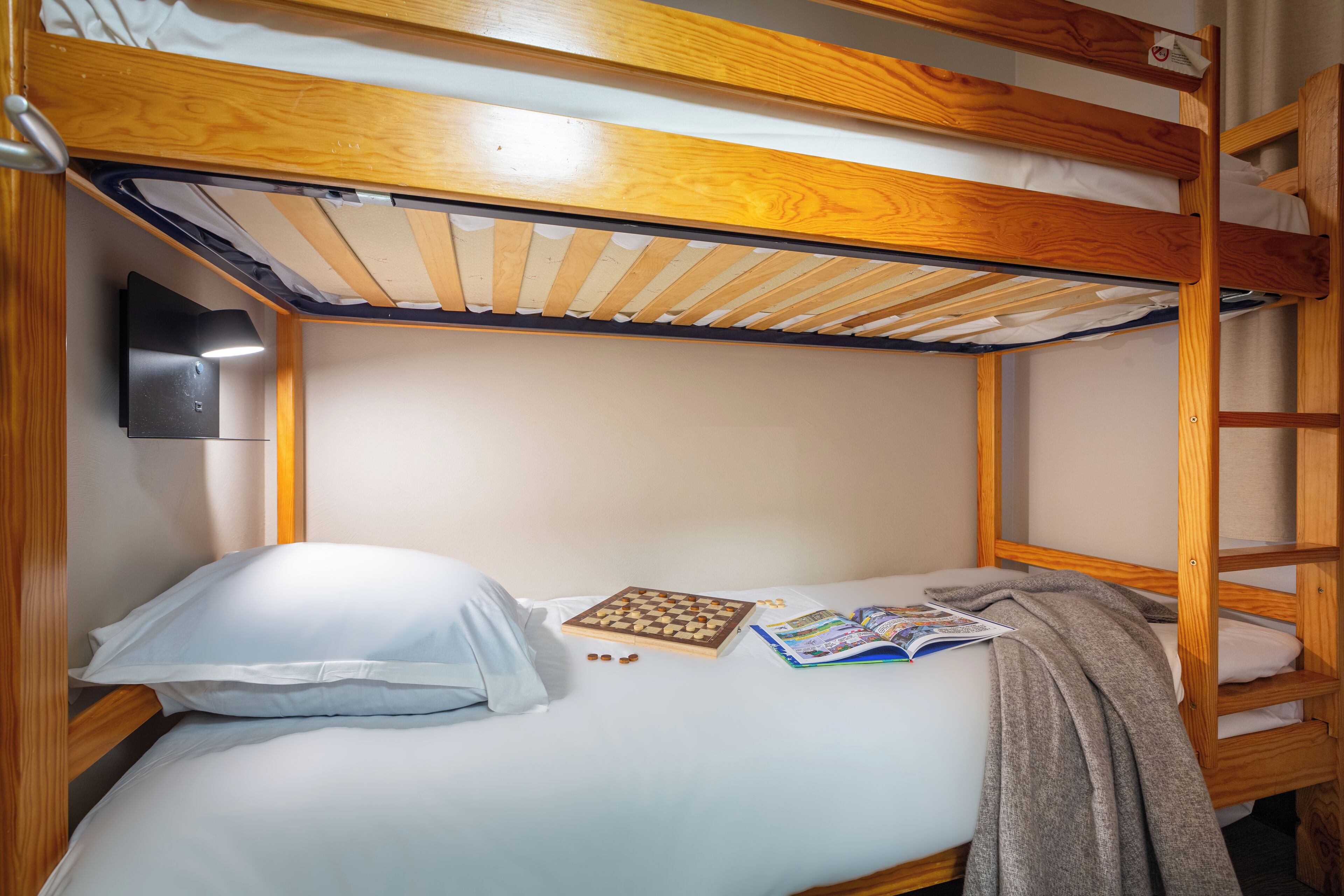 Una cama litera con estructura de madera, ropa de cama blanca, una almohada, un libro y una manta.