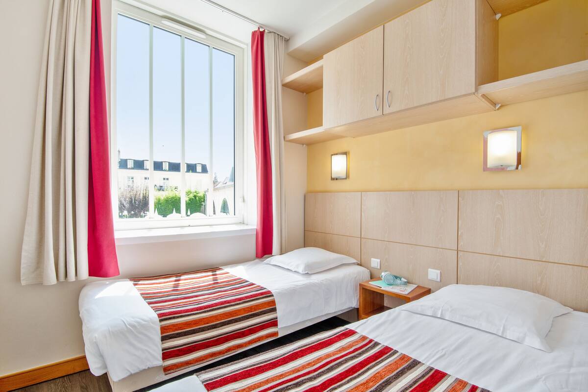 A hotel room with two beds, a window with curtains, a bedside table with a lamp, and a wall-mounted light.