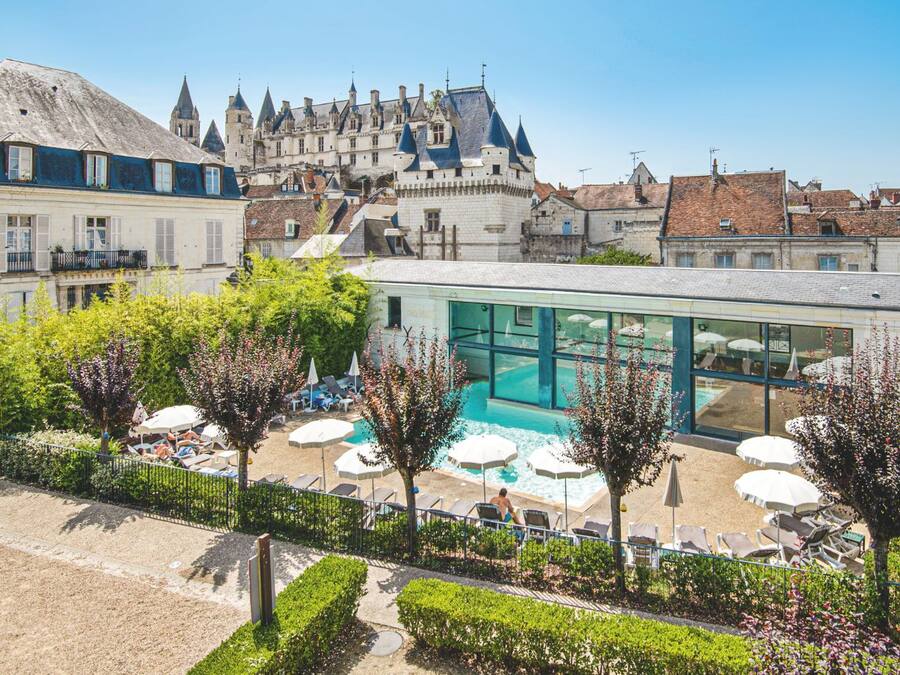 A hotel with a swimming pool, surrounded by lounge chairs and umbrellas, with a castle in the background.