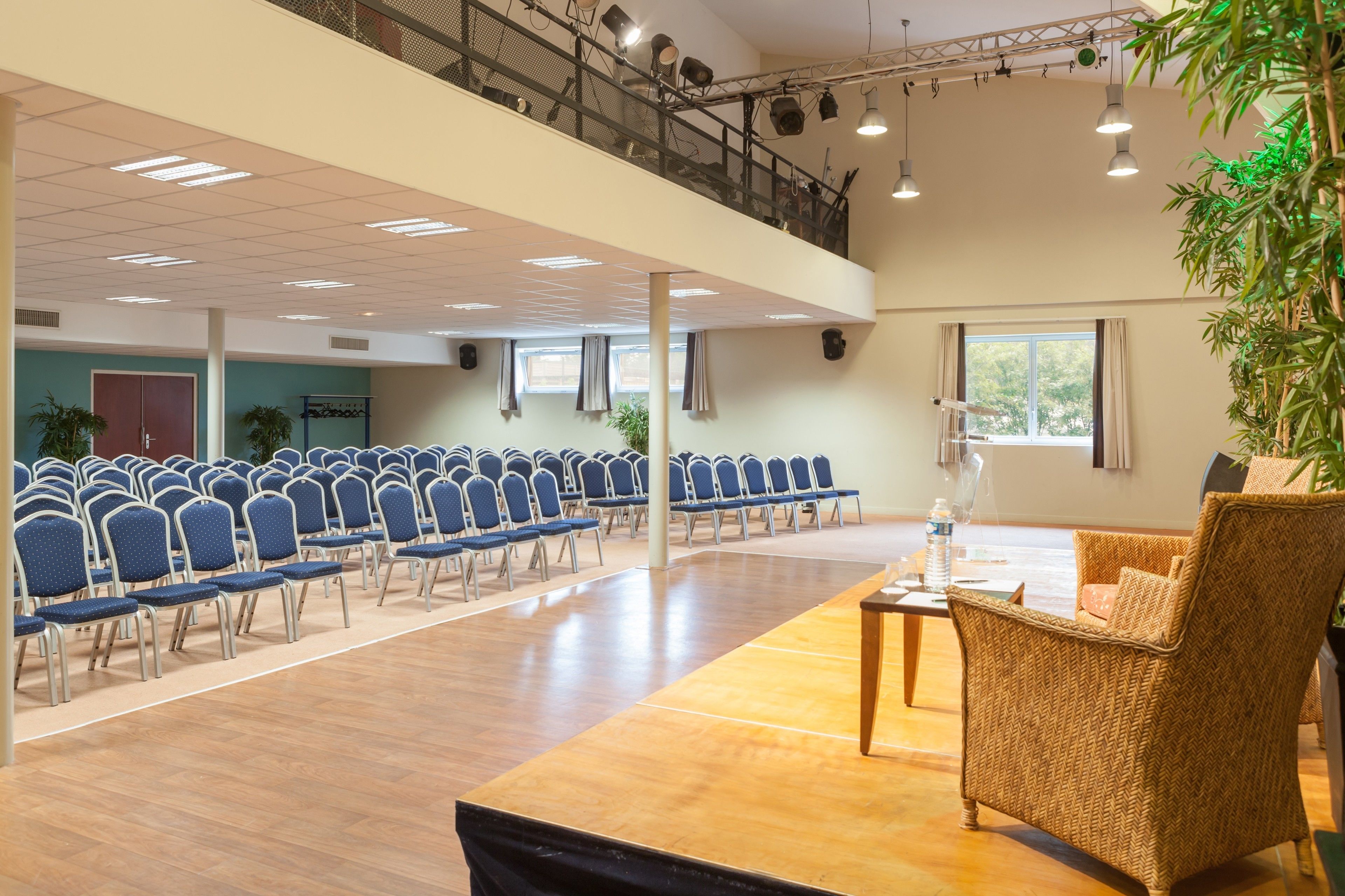 A spacious hall with rows of blue chairs, a wooden table, and a balcony with railings.