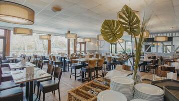 A spacious restaurant with multiple dining tables and chairs, large windows, and hanging pendant lights.