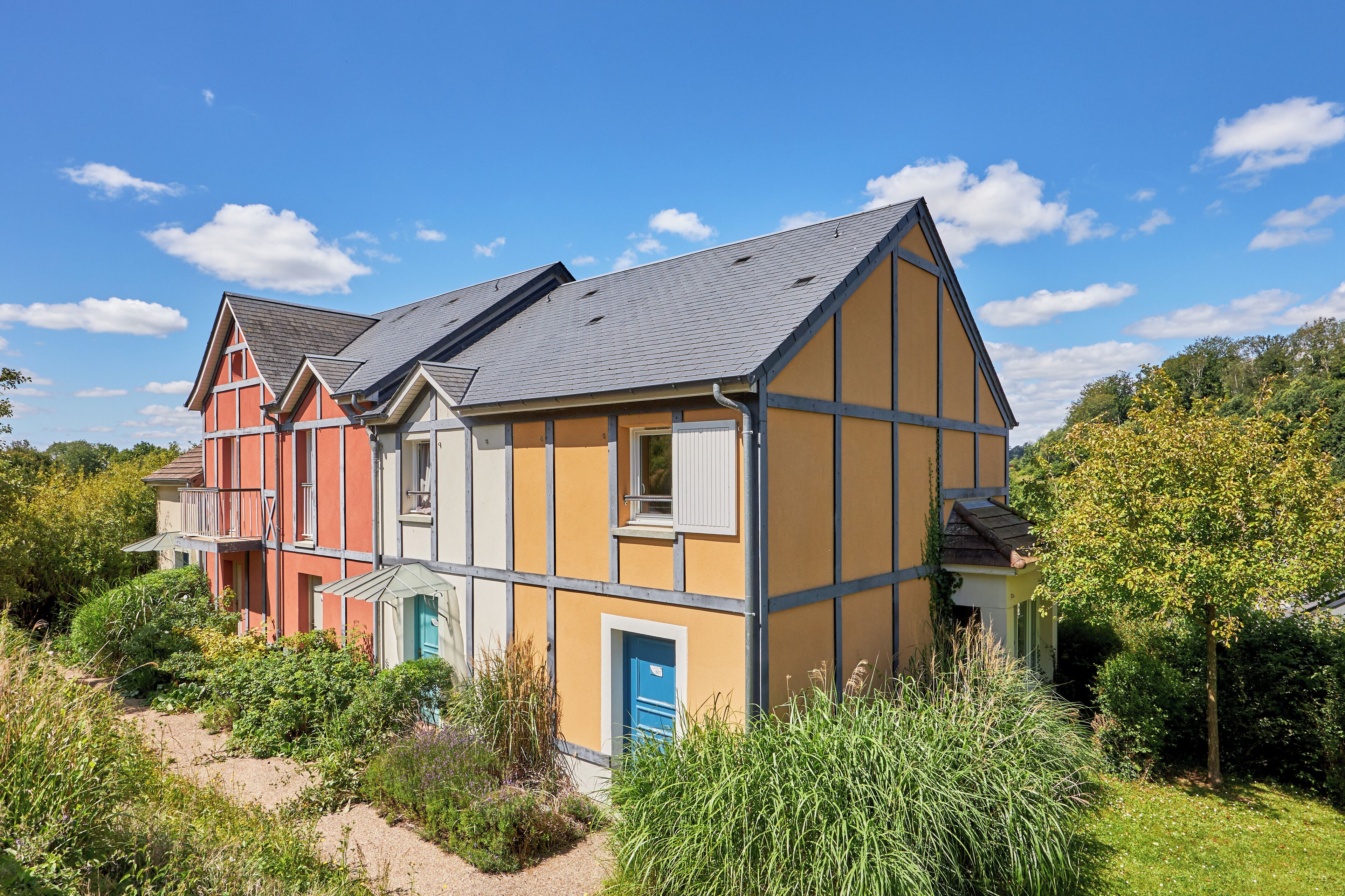 A multi-story house with a sloped roof, a balcony, and a garden area with various plants.