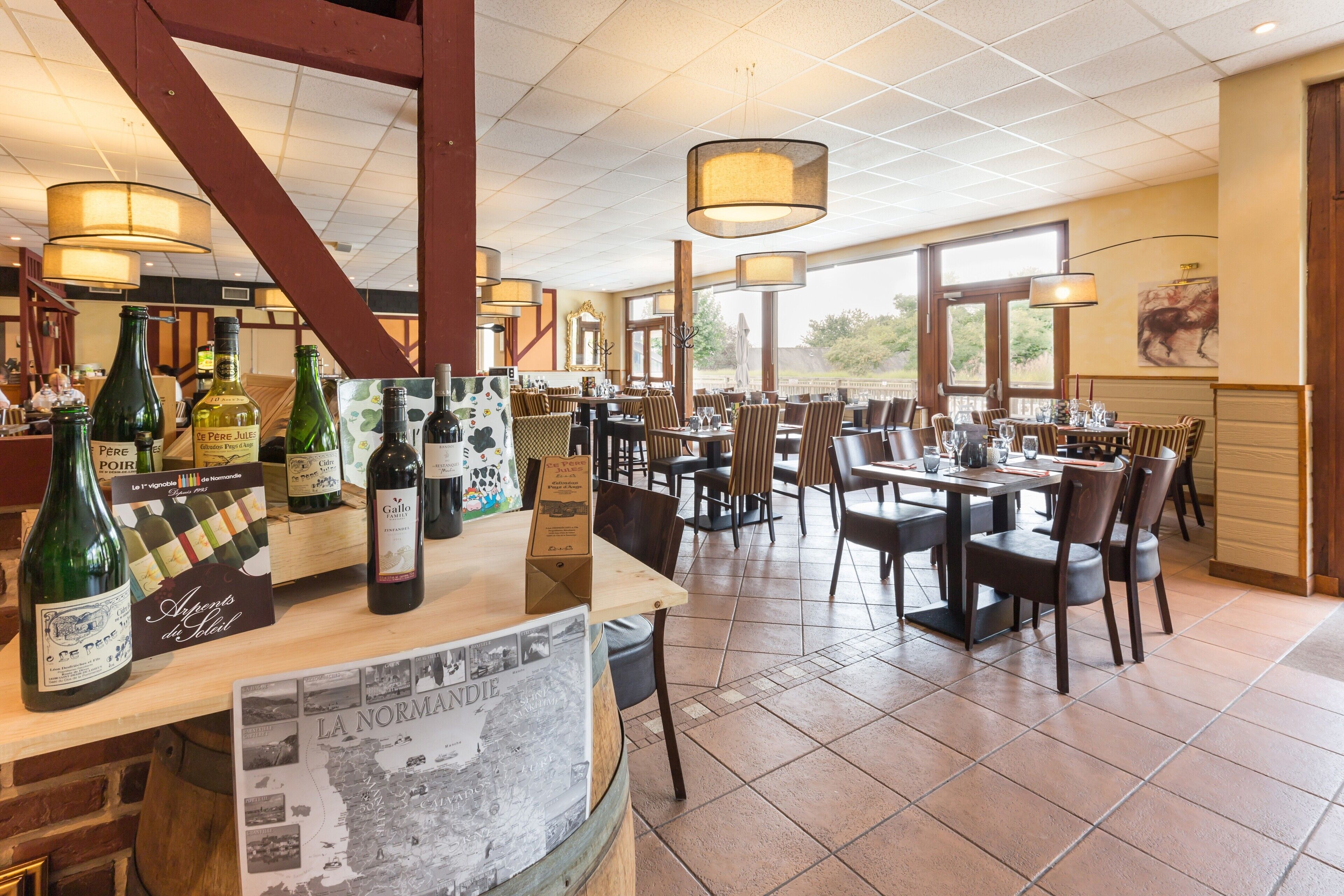 A restaurant interior with multiple dining tables and chairs, a wine display with various bottles, and a large window providing a view of the outdoors.
