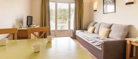 A room with a dining table, chairs, a sofa with patterned cushions, a TV, and a window with curtains.