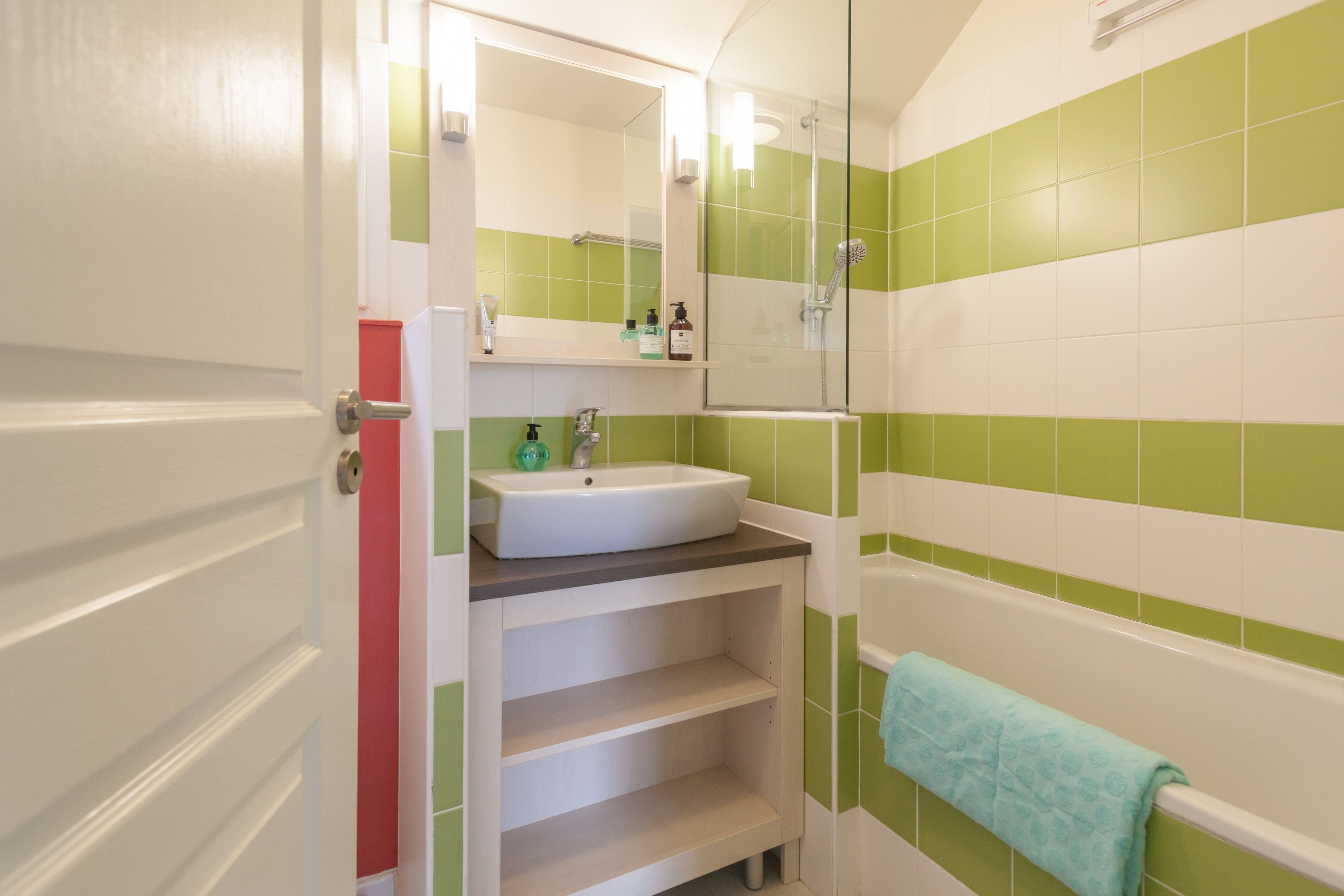 Un bagno con un lavandino bianco, piastrelle verdi e bianche, uno specchio e un asciugamano blu appeso alla vasca.