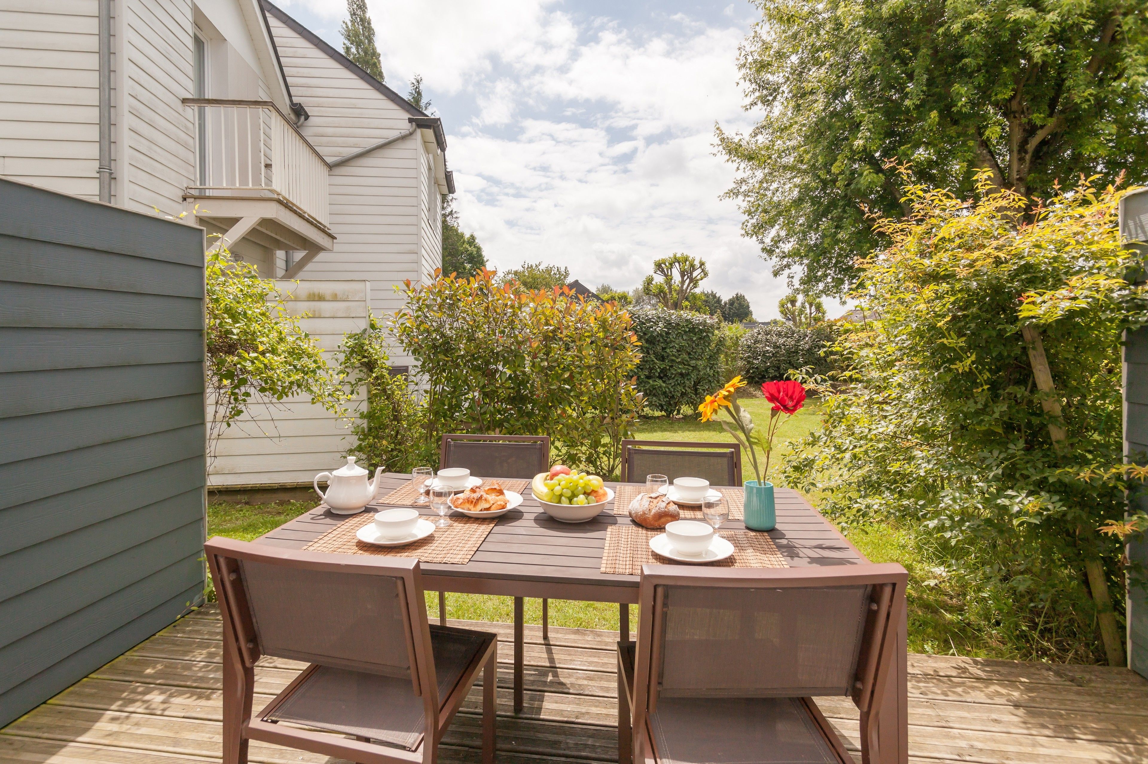 Een eethoek buiten met een tafel gedekt voor een maaltijd, omringd door stoelen, en uitzicht op een tuin met bomen en een huis op de achtergrond.