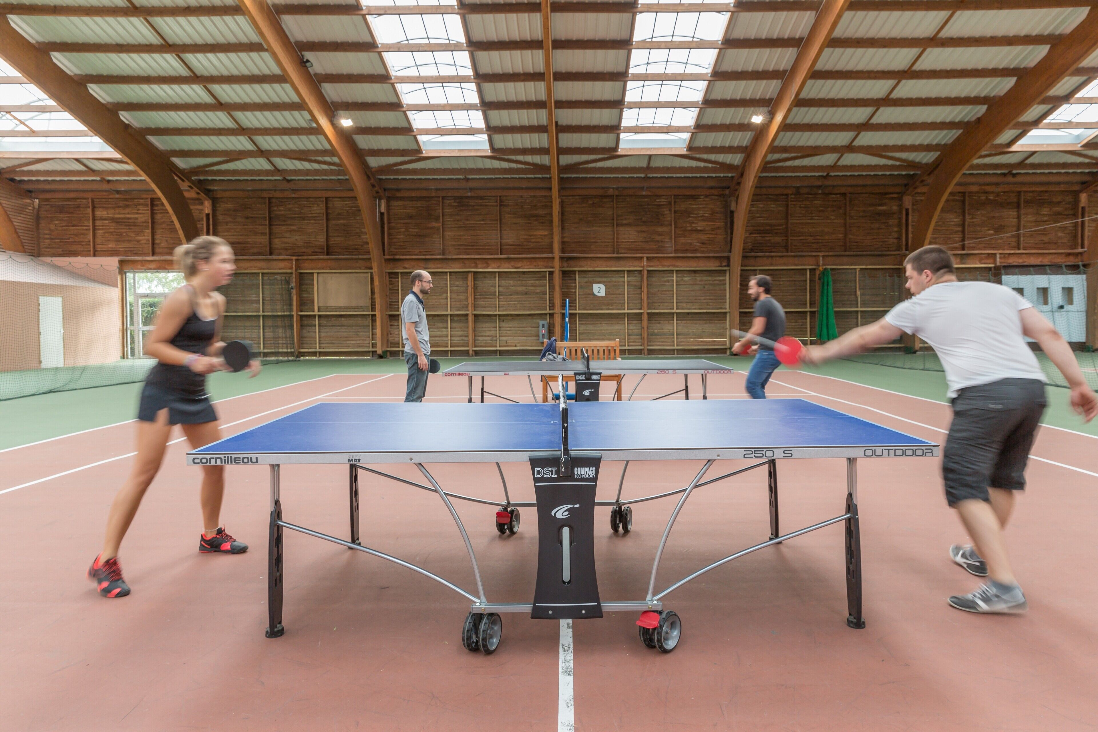 An indoor tennis court with a table tennis table, wooden walls, and a high ceiling with skylights.