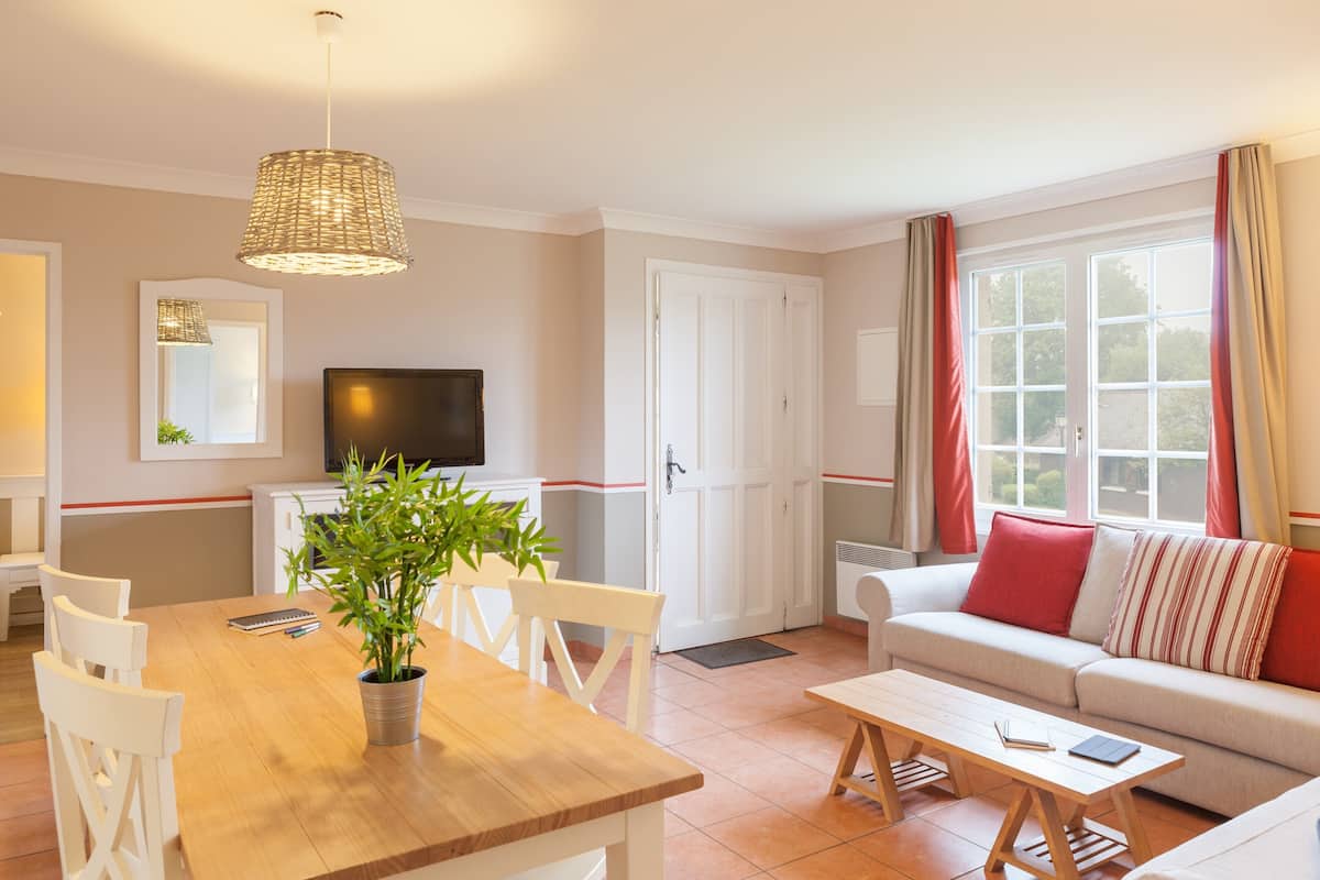 Een moderne woonkamer met een houten eettafel, witte stoelen, een bank met rode kussens, een televisie en een groot raam met gordijnen.