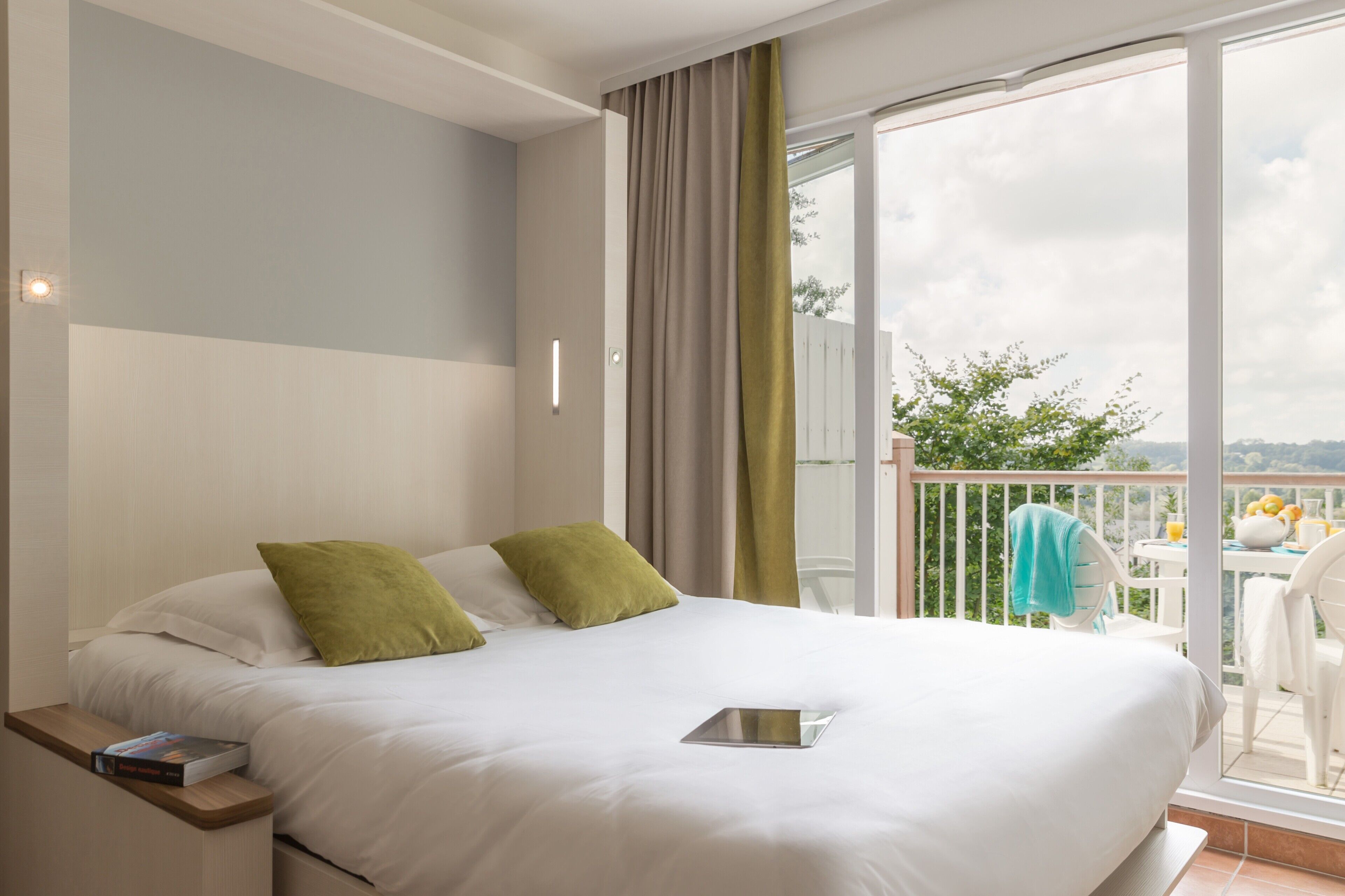 A hotel room with a bed, a bedside table with a book, a tablet, and a balcony with chairs and a table.