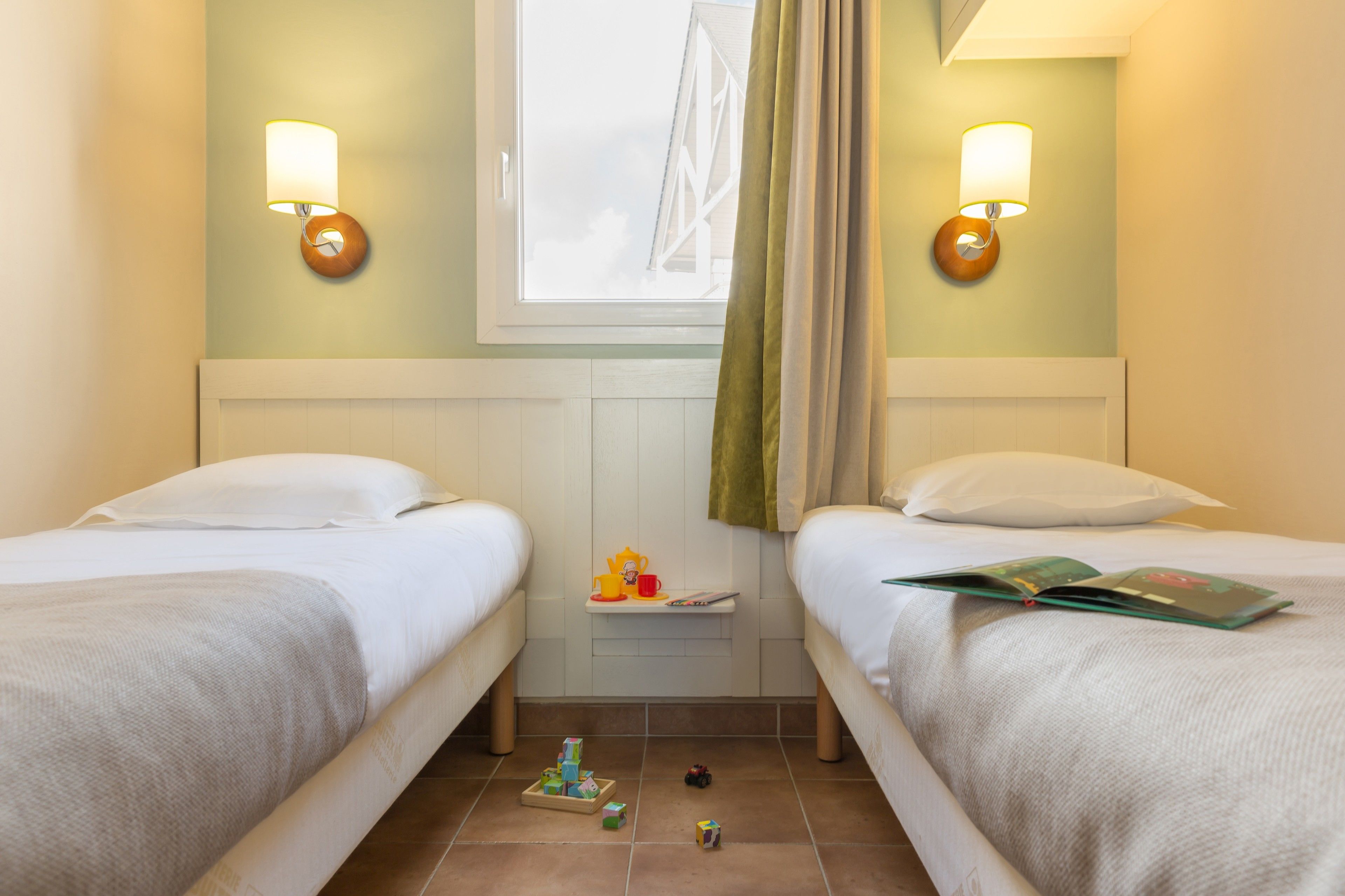A hotel room with two beds, a small table with toys, and wall-mounted lamps.