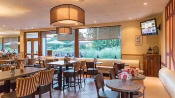 A restaurant with multiple dining areas, including booths and tables with striped chairs. There are large windows with views of greenery outside.