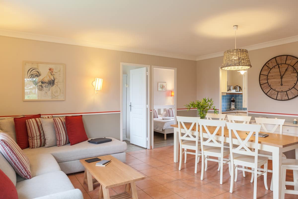 Een woonkamer met een bank, een eettafel en stoelen, een keuken op de achtergrond en een grote wandklok.